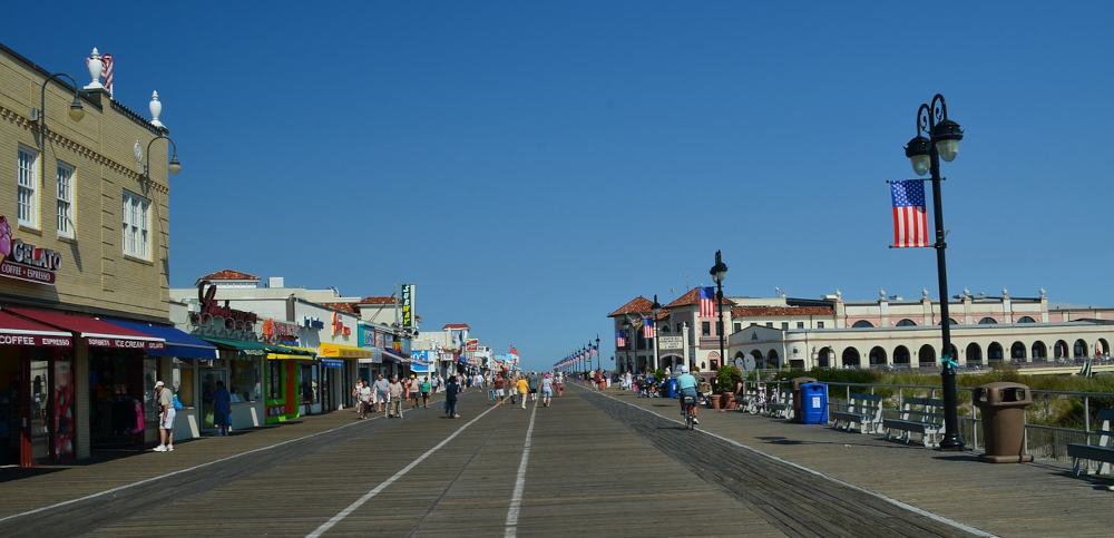 NJ Atlantic City Boardwalk is seen on a sunny day with blue skies. A street stretches into the distance lined with shops and casinos. to the right is the beach. AC is an iconic gambling city, famous for its casinos. NJ Online Casinos are also very popular
