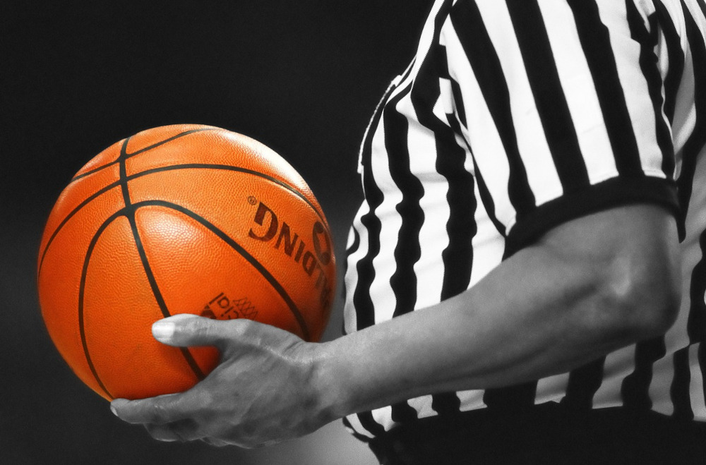 the midsection of a basketball ref is seen, holding a basketball in front of him. everything is in black and white except for the ball, which is bright orange.
